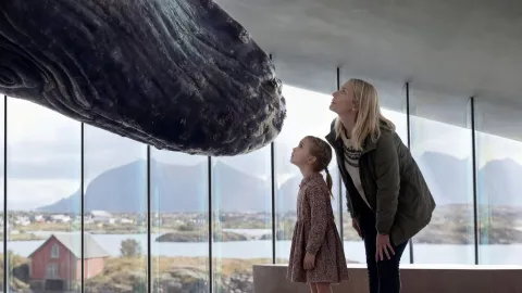 Two visitors stand by large windows beneath a suspended whale, with water, hills, and a distant building outside.