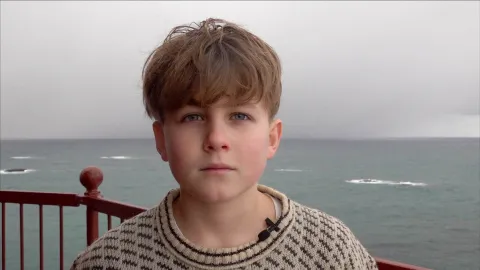 Person in a patterned sweater stands on a balcony with red railing, ocean waves and an overcast sky behind.