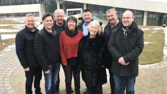 A group of adults stands on a paved path outdoors near a modern glass building with patches of snow on grass.