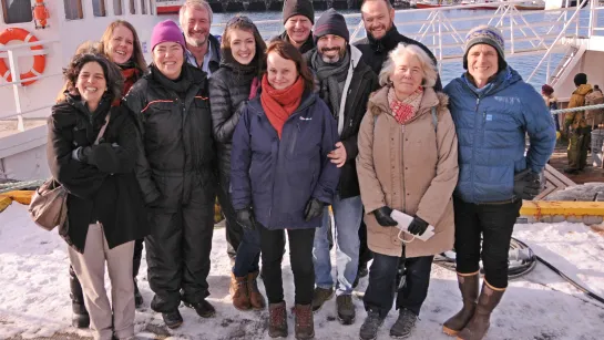 Group in winter coats and boots standing on a snowy dock beside a boat, with harbor water and buildings behind.