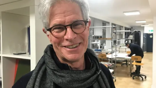 Indoor workshop scene with a person wearing a dark scarf in the foreground, shelves and worktables behind.