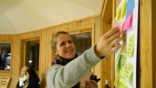 A person in a sweater points at a wall board covered with colorful sticky notes inside a wooden room.