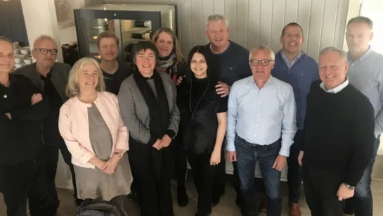 A group of adults standing together indoors in casual and smart-casual clothing, with a cabinet behind them.