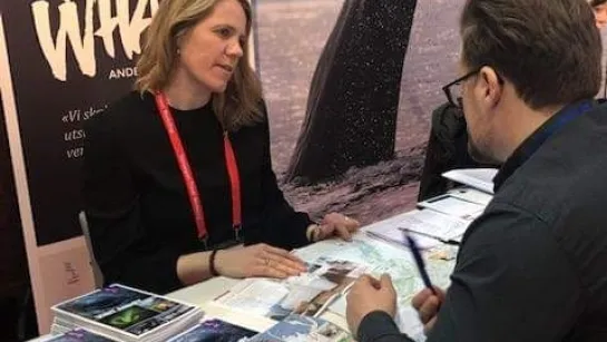 Two people sit at a travel information booth with maps and brochures on a table and a large whale poster behind.