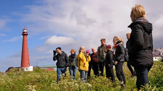 A group of people stands in a wildflower field near a tall red lighthouse under a partly cloudy sky.