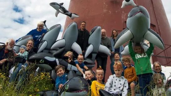 Low-angle view of a tall red lighthouse with many inflatable whales and sharks piled below under a cloudy sky.