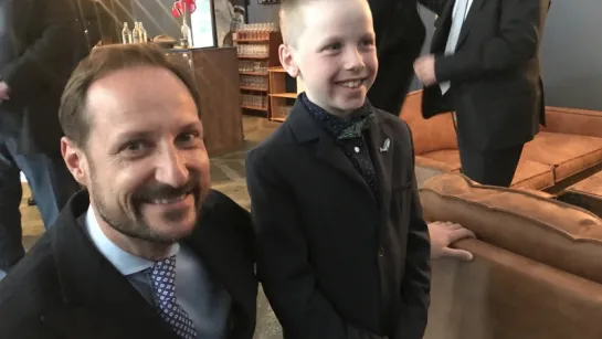 Two well-dressed guests in suits stand in a lounge area with leather sofas, people, and a bar in the background.