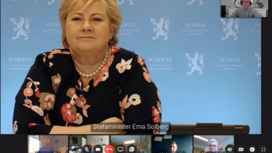 Person in floral dress and pearl necklace sits before a blue Norway government backdrop during a video conference.