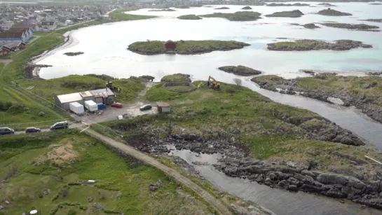 Aerial view of rocky coastline with grassy land, small islands, a lagoon, and a roadside worksite with vehicles.