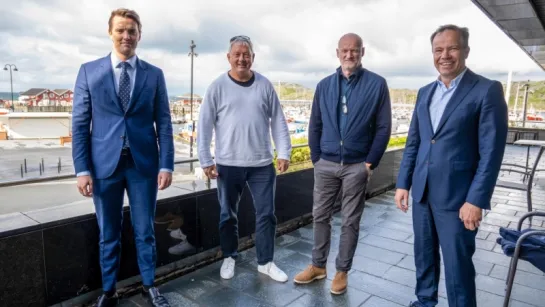 Four men stand on a tiled terrace by a marina, with boats, buildings, and cloudy sky in the background.