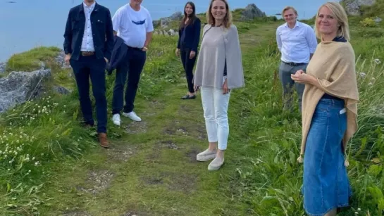 Five adults stand on a grassy coastal trail among rocks and wildflowers, with ocean and cloudy sky behind.
