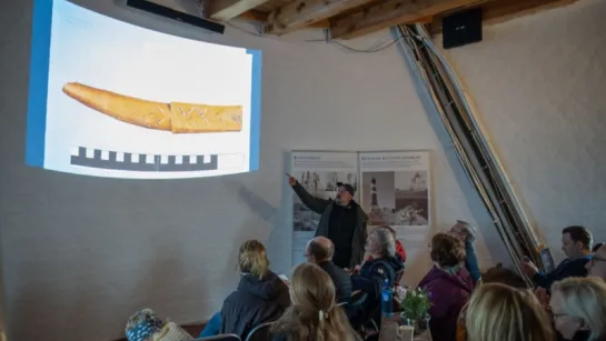 A guide gestures toward a projected photo of an artifact in a small room as seated visitors listen.