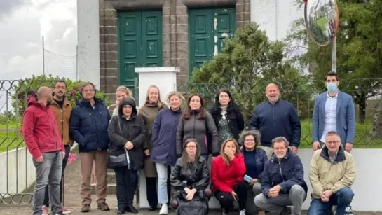 A group of adults posing outdoors on steps in front of a white building with green doors and a fence.