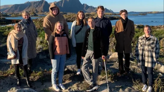 A group stands on rocky ground by the sea with mountains in the background under clear blue sky.