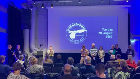 People sit in an auditorium facing a stage where a group stands beside a large projected screen with a whale logo.