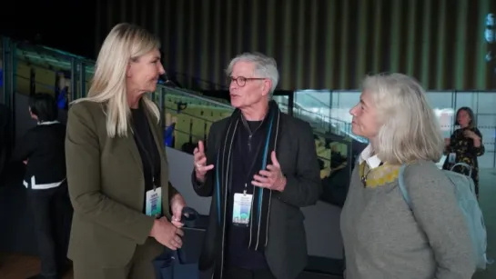 Three attendees wearing lanyard badges talk in a modern indoor venue with seating, railings, and overhead lights.