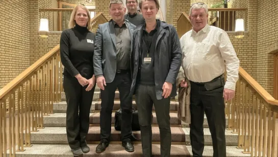 Four adults stand on a wide staircase inside a brick-walled atrium with railings, lamps, and a window above.
