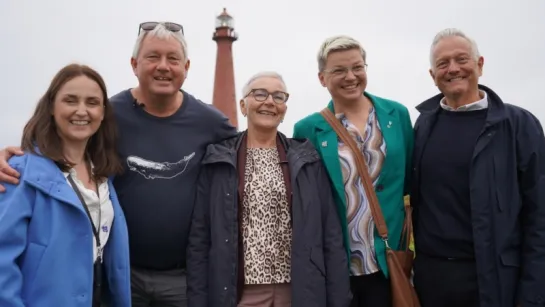 Five adults stand together outdoors with arms around each other, with a tall lighthouse in the background.