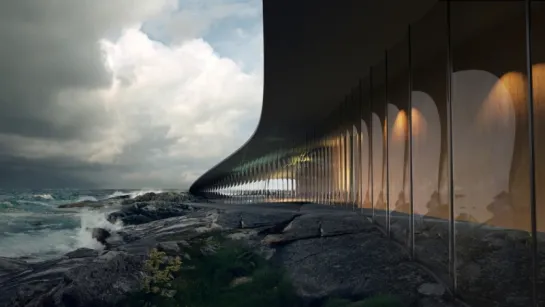 Curving modern glass building along rocky shore, with waves crashing under a cloudy sky and warm interior lights.
