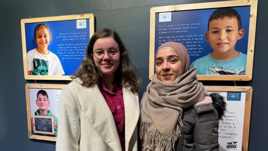 Two people stand indoors in front of framed portrait panels with handwritten text on a dark blue wall.