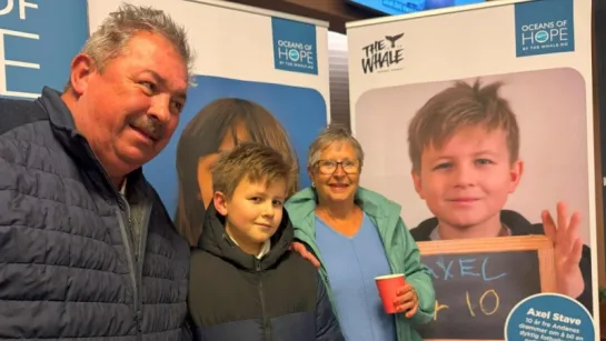 Three people stand indoors in front of blue-and-white informational posters; one holds a red cup.