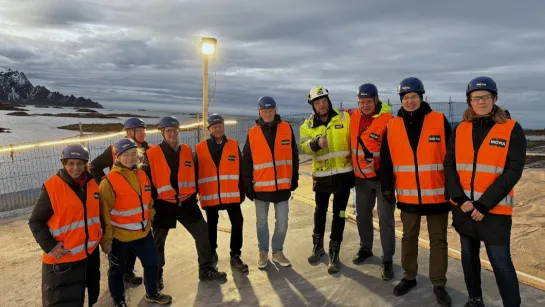 Group in orange safety vests and helmets standing on a platform near a fenced coastline with mountains under clouds.