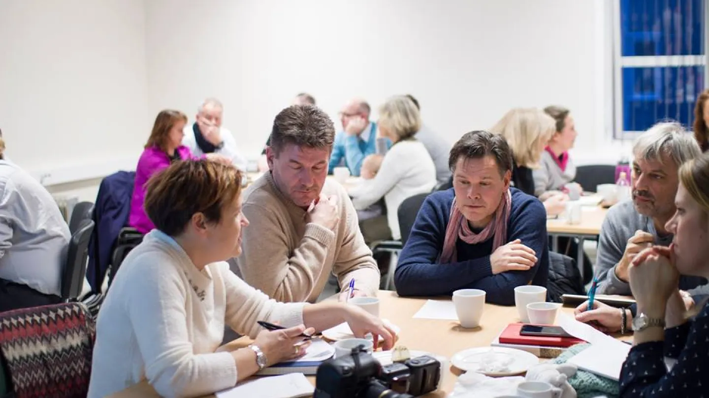 People sit around a table in a meeting room, talking and taking notes with coffee cups and a camera on the table.