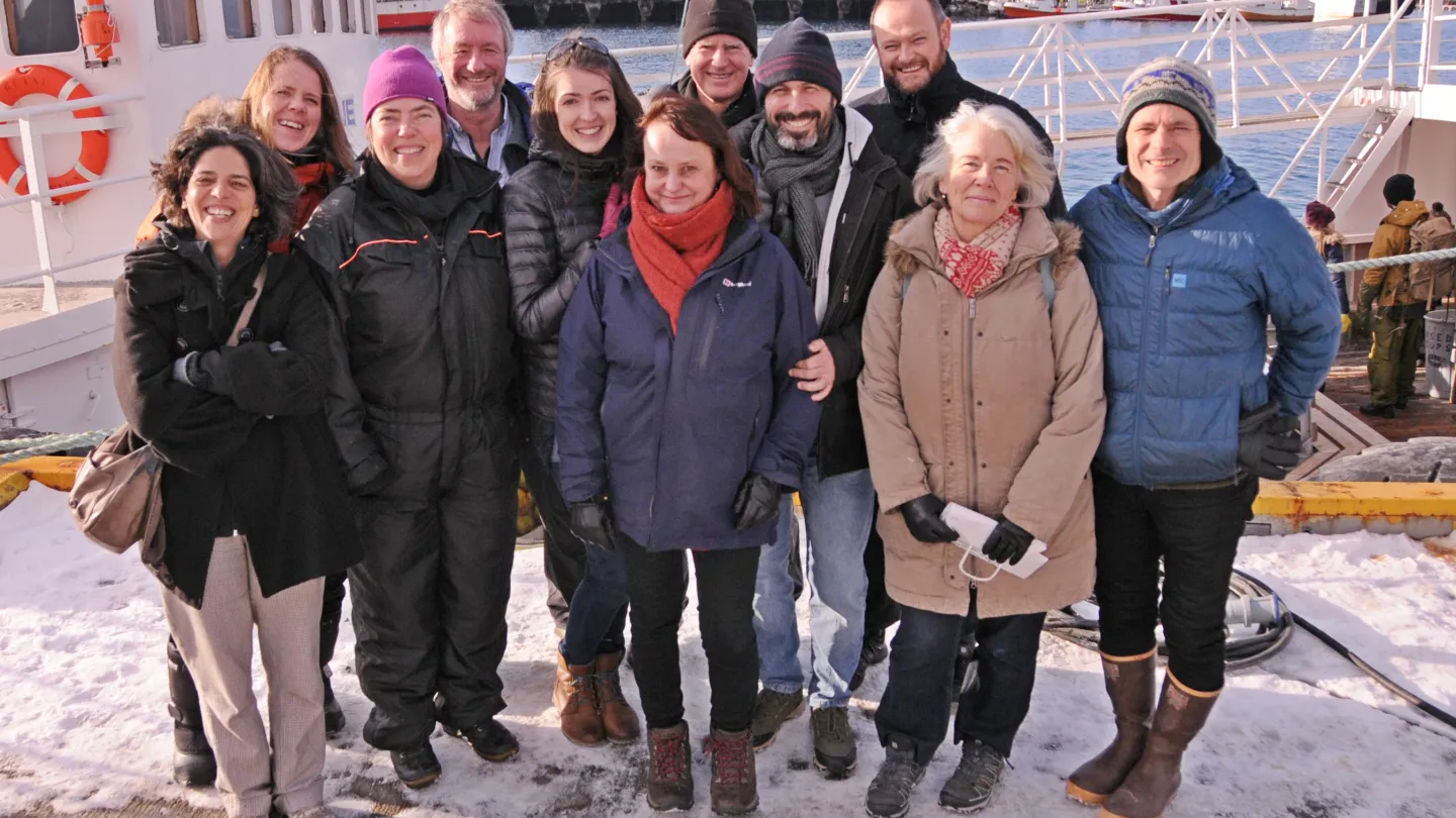 Group in winter coats and boots standing on a snowy dock beside a boat, with harbor water and buildings behind.