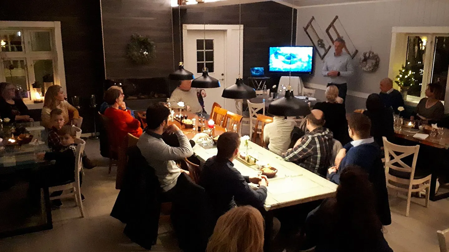 Group seated at long tables in a warmly lit room, watching a standing speaker beside a TV screen and decor.