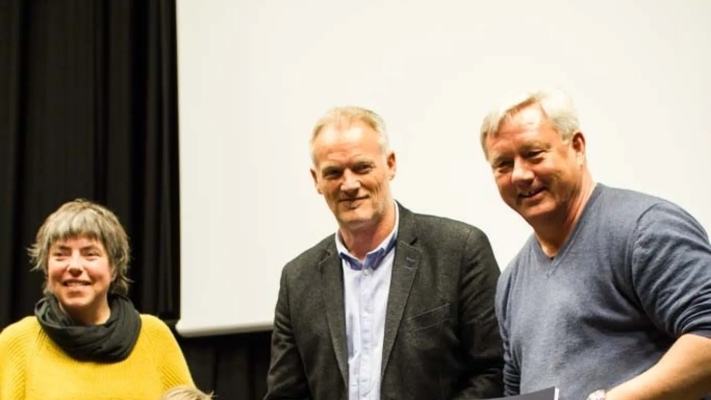 A group stands indoors on a stage area, wearing white gloves and holding large spiral-bound booklets.