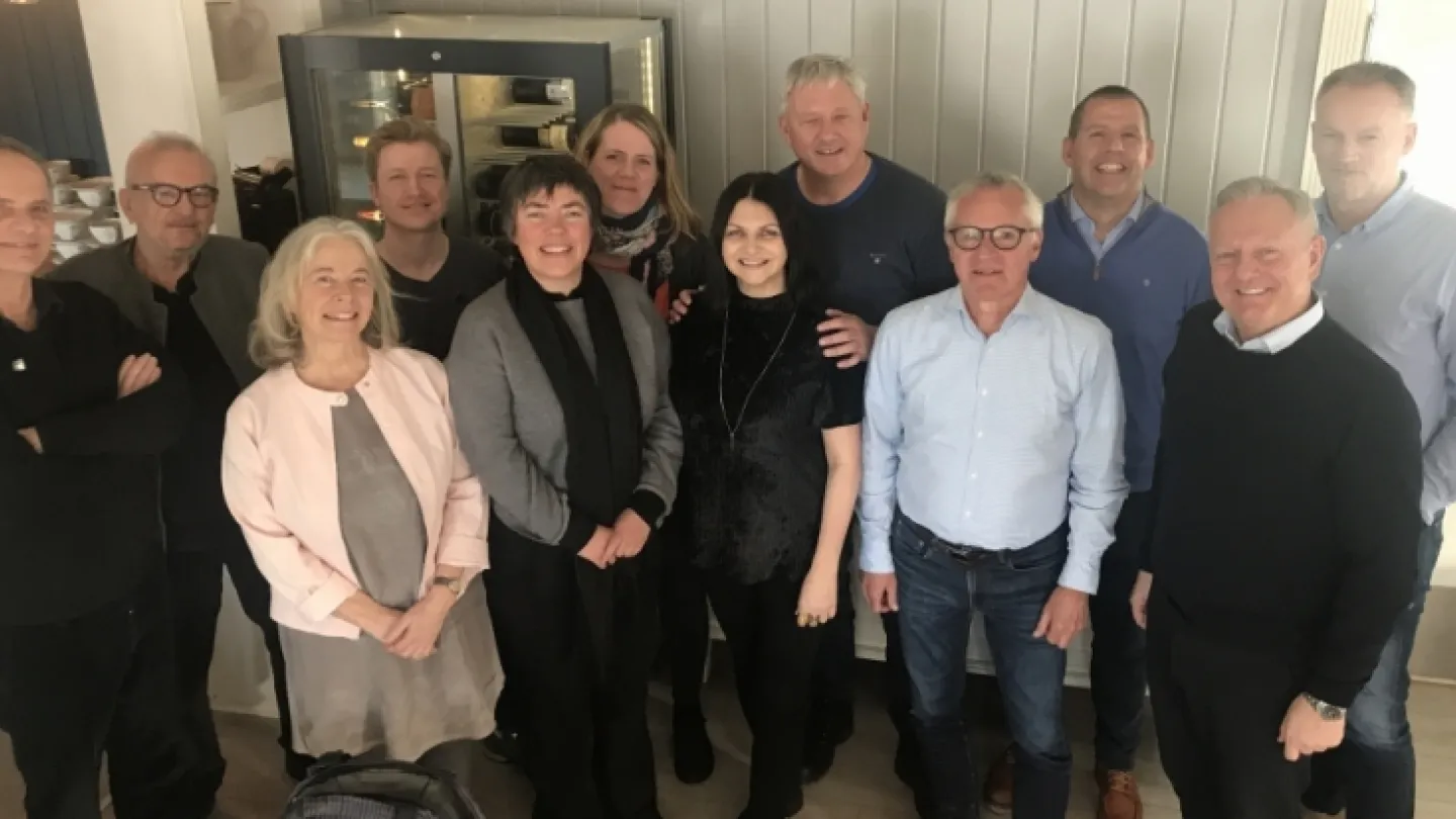 A group of adults standing together indoors in casual and smart-casual clothing, with a cabinet behind them.
