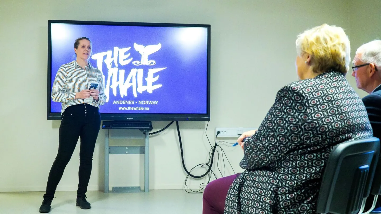 A person stands beside a large TV displaying “THE WHALE” while two attendees sit in chairs in a plain room.