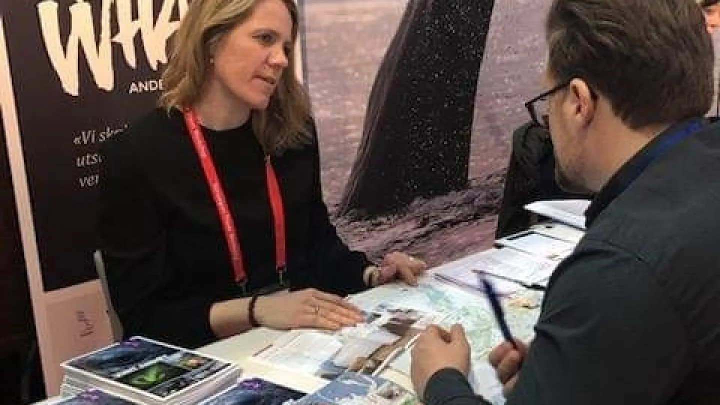 Two people sit at a travel information booth with maps and brochures on a table and a large whale poster behind.