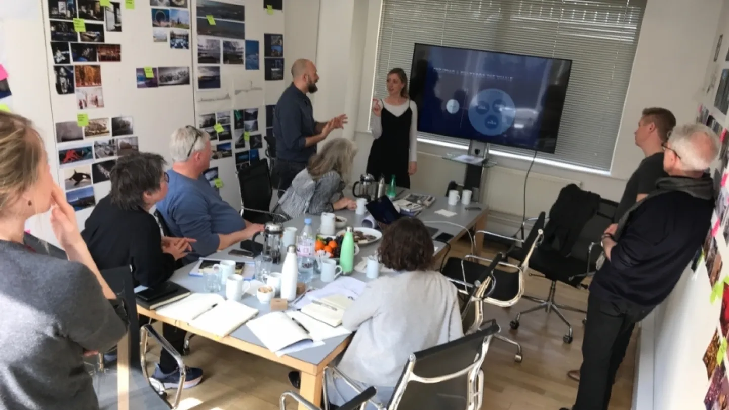 Group gathered around a conference table in an office, discussing ideas while a presenter stands near a large screen.