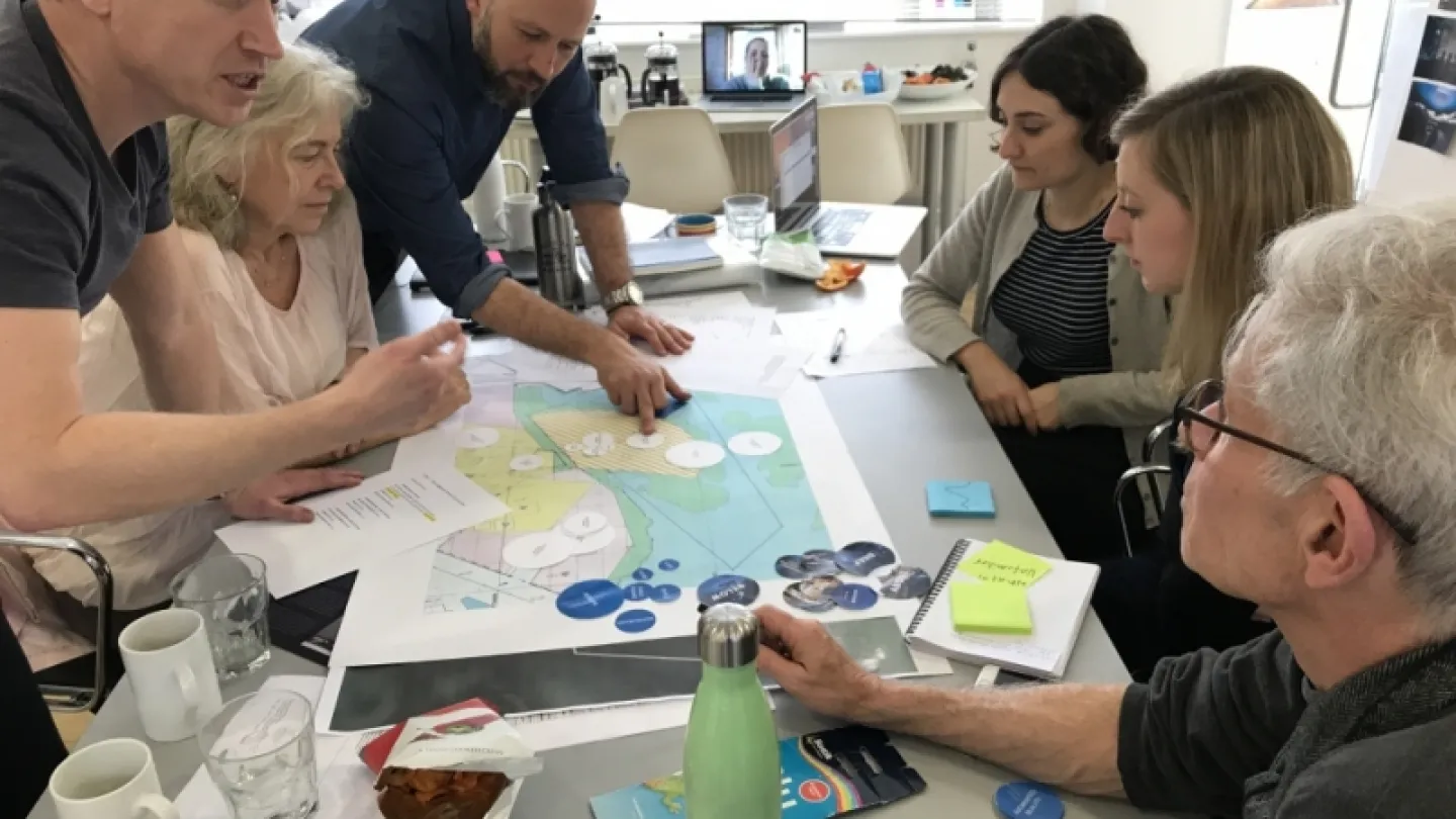 Group gathered around a table reviewing a large map, papers, and notes in a bright office meeting space.
