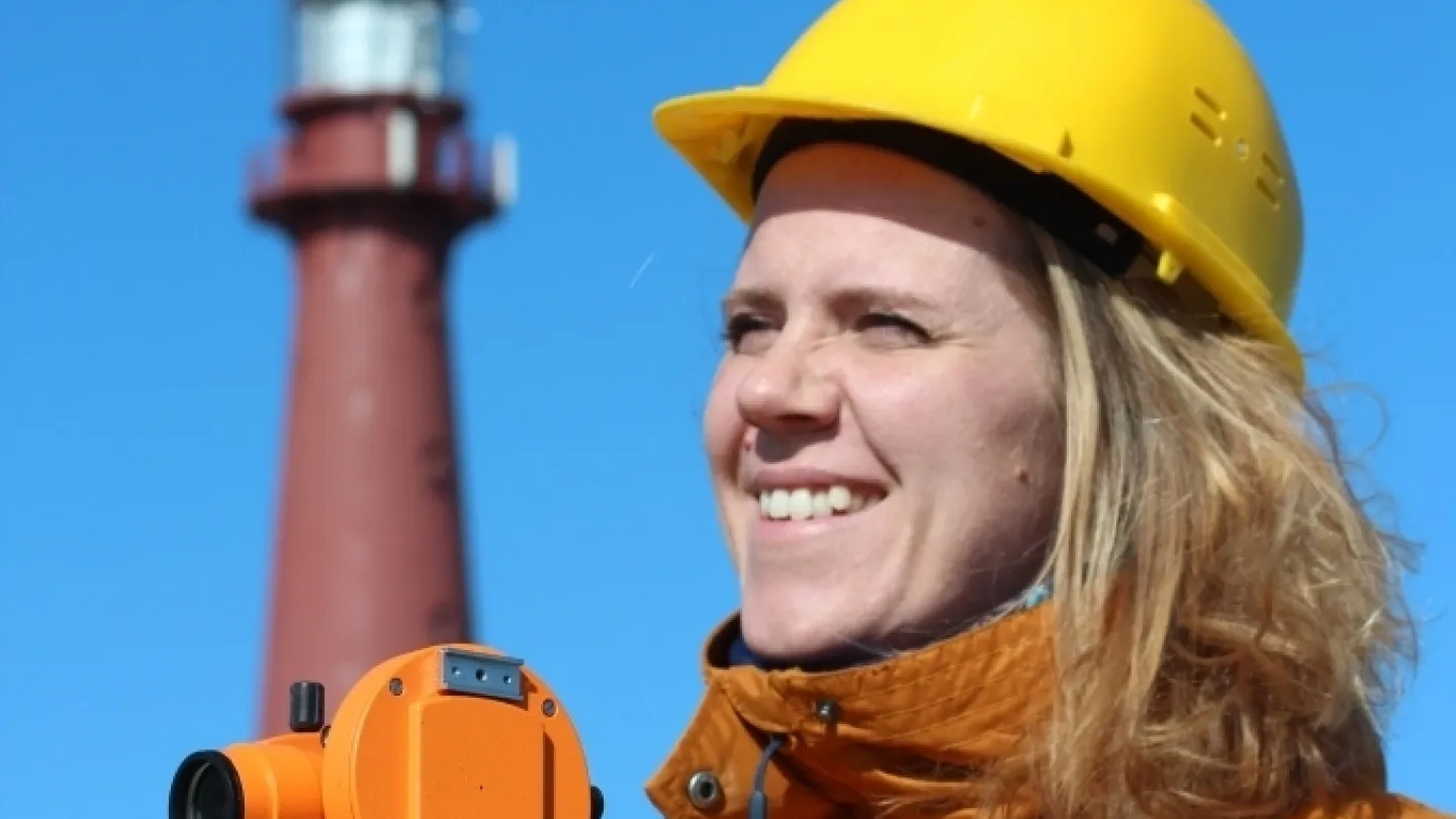 Person in a yellow hard hat and orange jacket adjusts a surveying instrument on a tripod with a lighthouse behind.