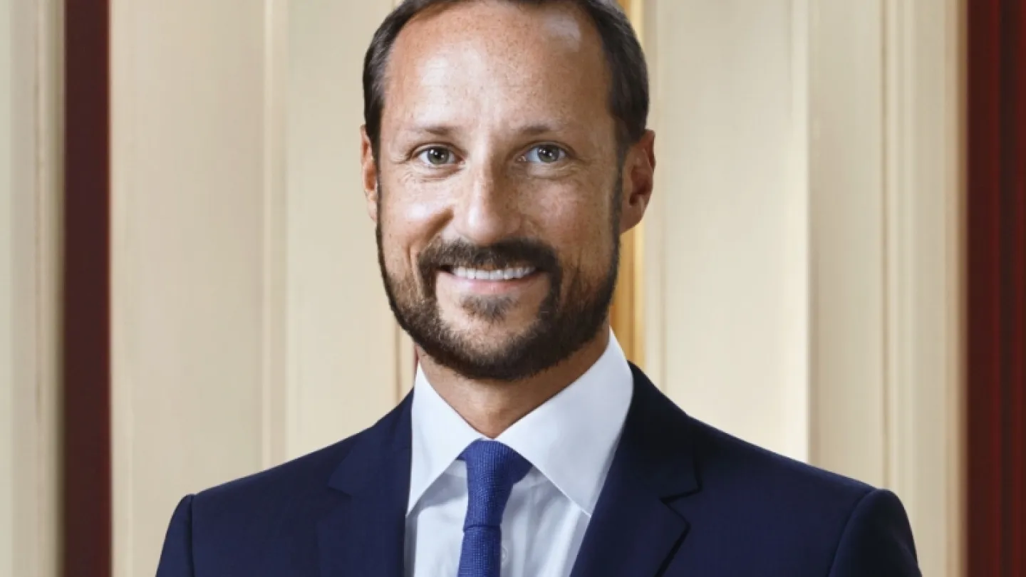 A person in a dark suit with a blue tie and white shirt stands indoors against cream and wood-paneled walls.