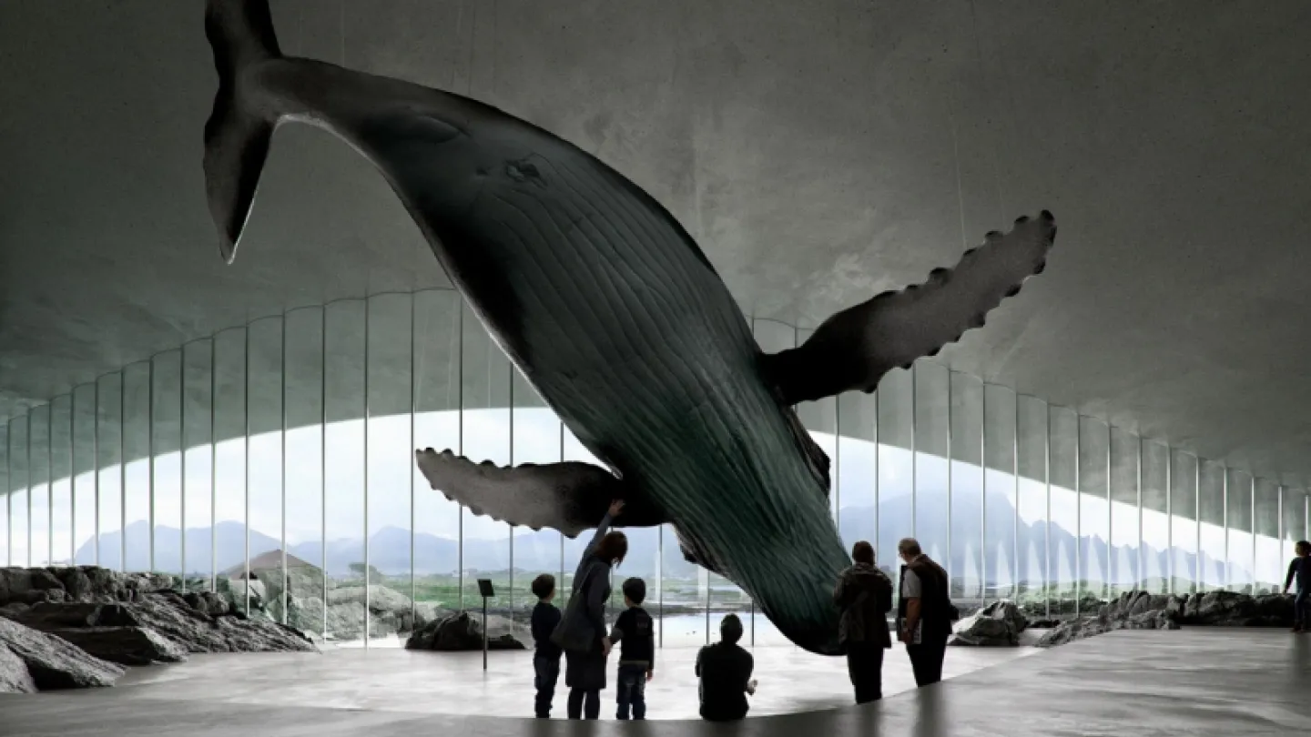 Large whale sculpture hangs above visitors inside a modern pavilion, with arched windows showing water and mountains.