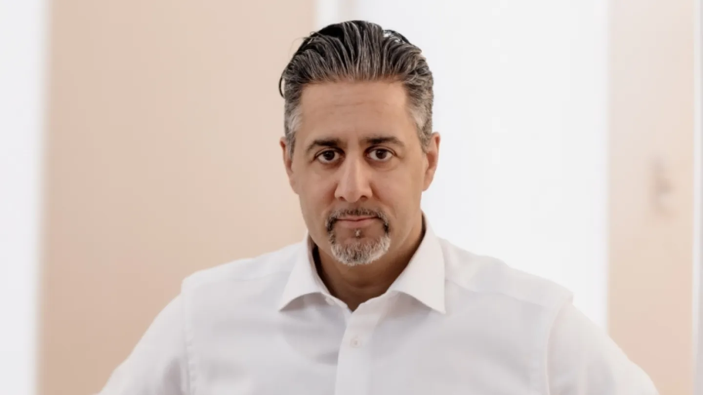 Upper-body view of a man wearing a white button-up shirt indoors, with a softly lit neutral background.