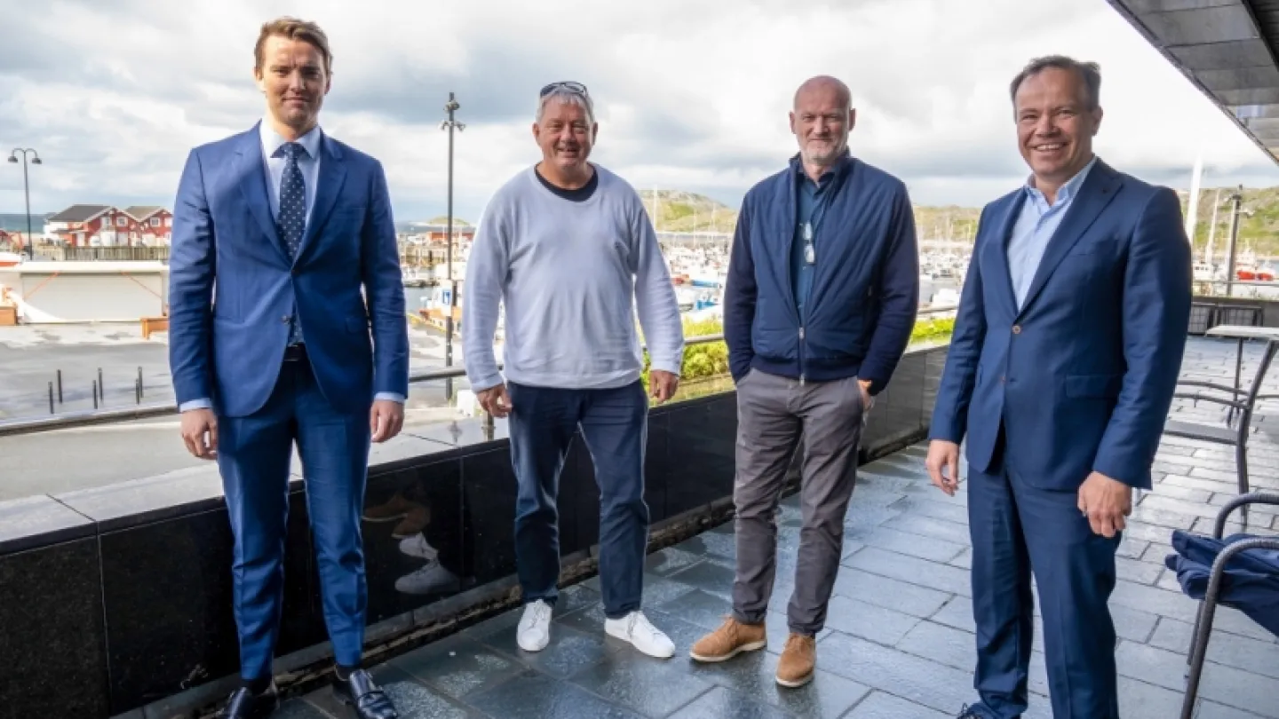 Four men stand on a tiled terrace by a marina, with boats, buildings, and cloudy sky in the background.
