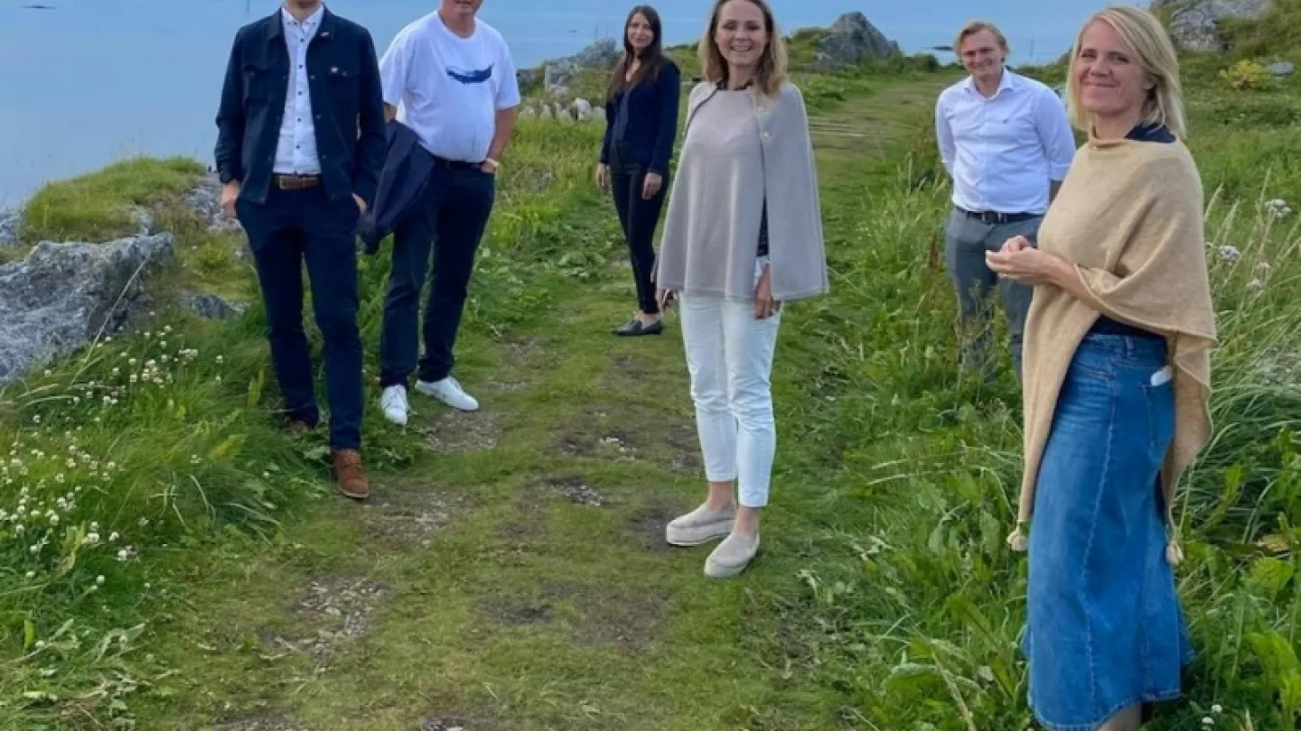 Five adults stand on a grassy coastal trail among rocks and wildflowers, with ocean and cloudy sky behind.