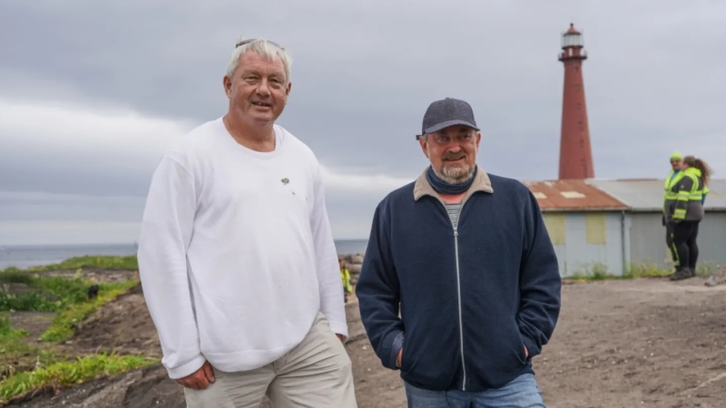 Two people stand on a rocky shore near a red lighthouse and small building under an overcast sky.