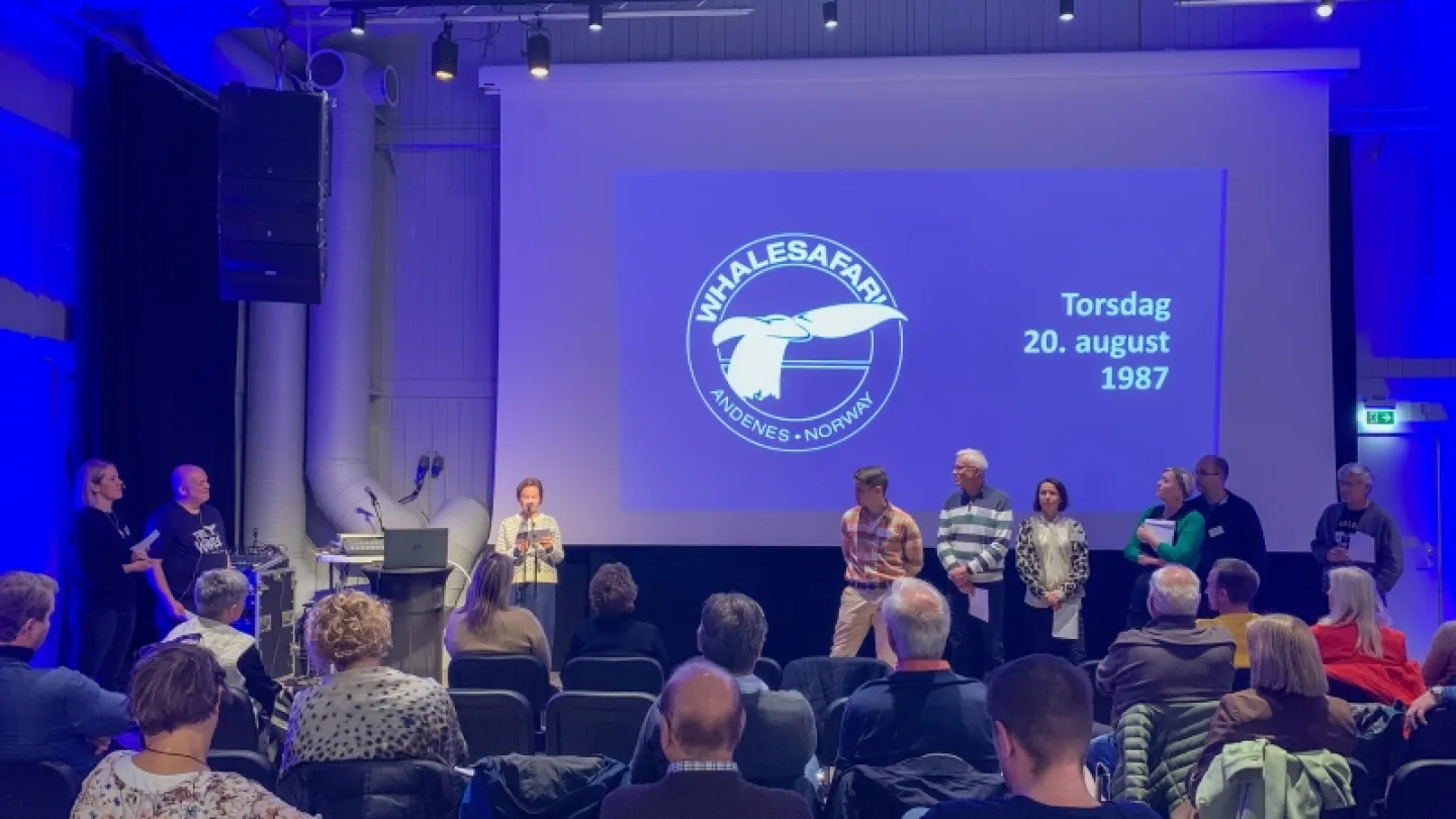 People sit in an auditorium facing a stage where a group stands beside a large projected screen with a whale logo.