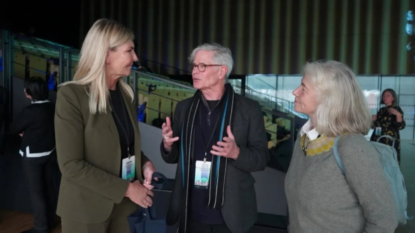 Three attendees wearing lanyard badges talk in a modern indoor venue with seating, railings, and overhead lights.