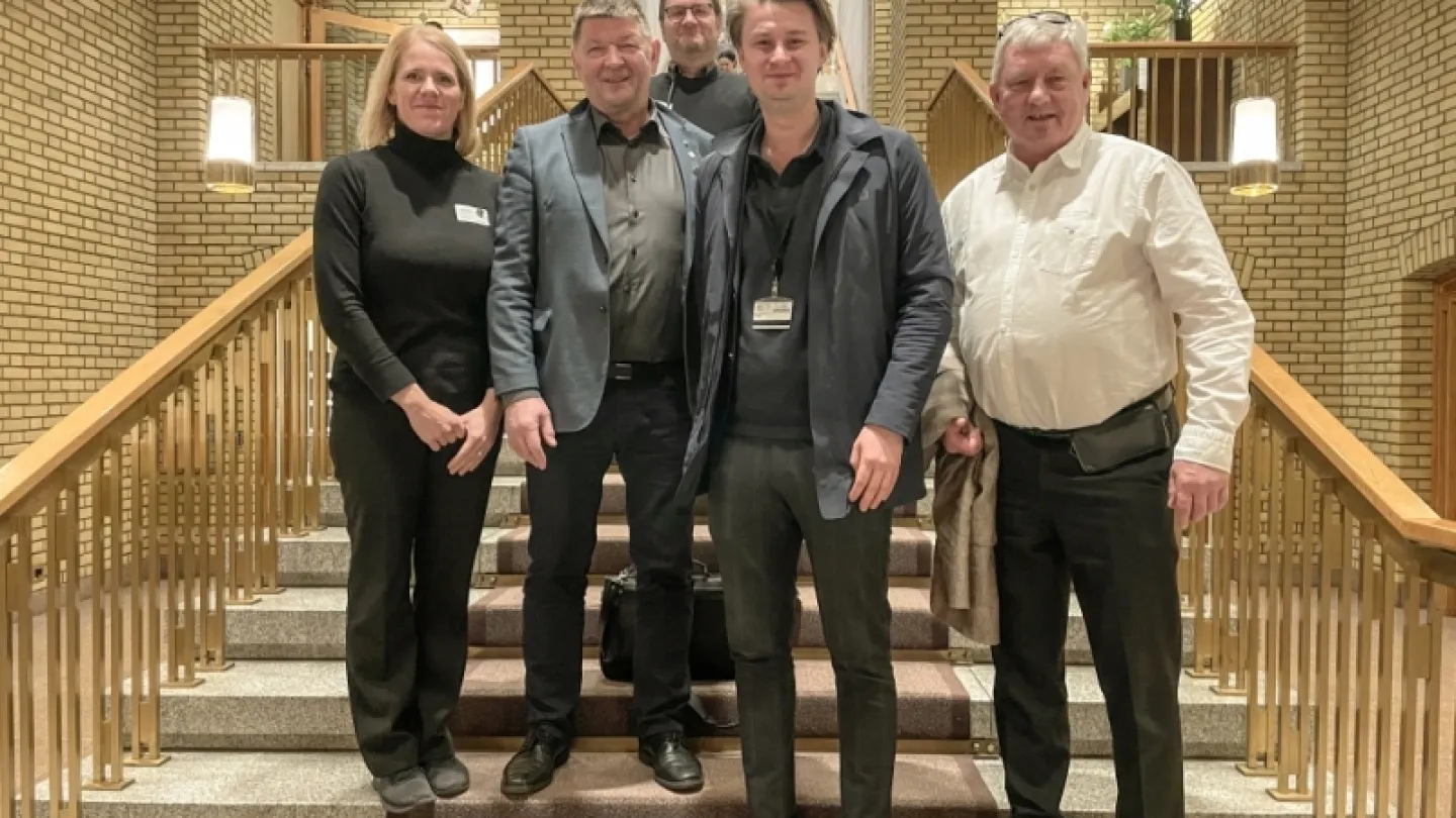 Four adults stand on a wide staircase inside a brick-walled atrium with railings, lamps, and a window above.