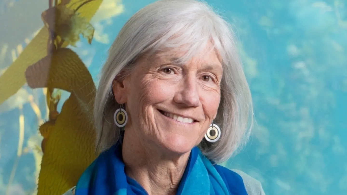 Seated adult with gray hair wearing a bright blue scarf and gray top, arms crossed, against leafy blue backdrop.