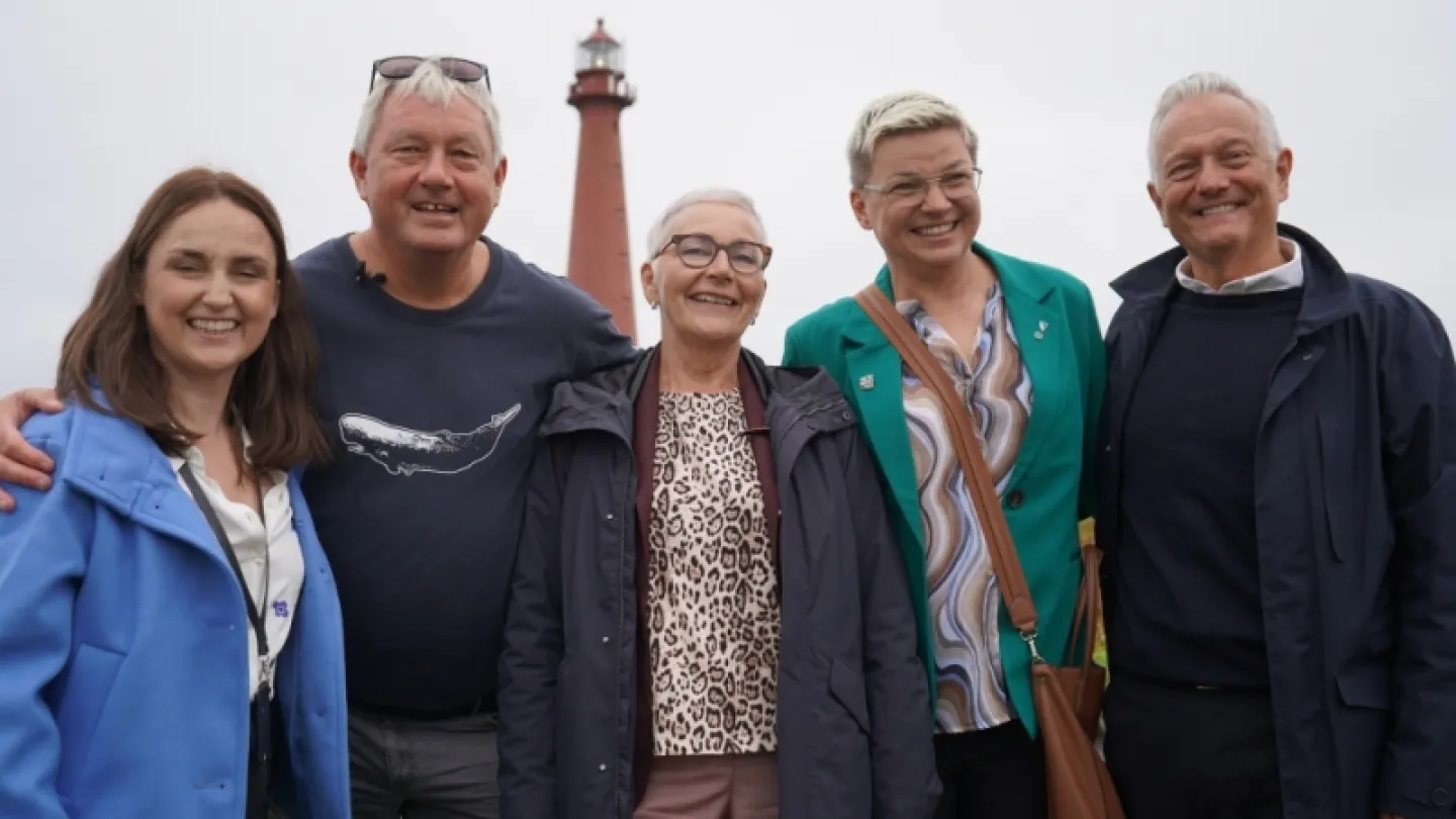 Five adults stand together outdoors with arms around each other, with a tall lighthouse in the background.