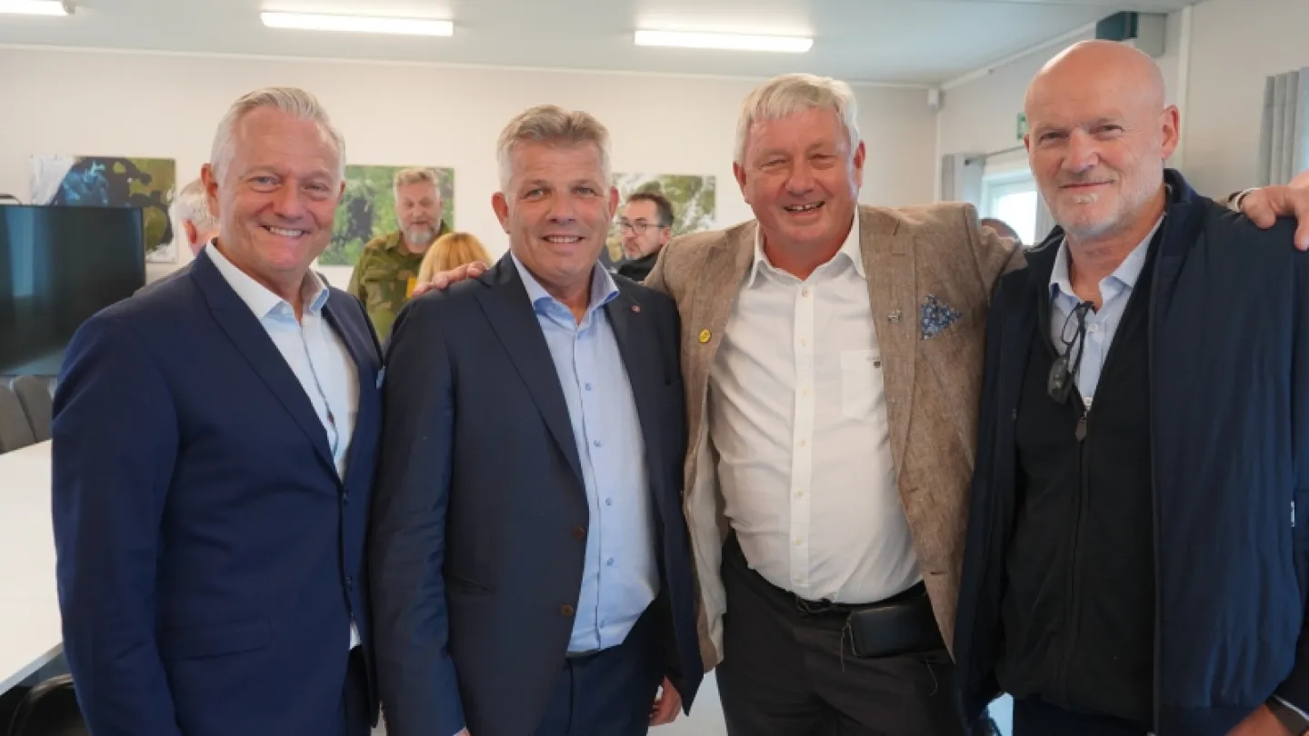 Four men stand close together with arms around shoulders in a bright office room with tables and wall art.