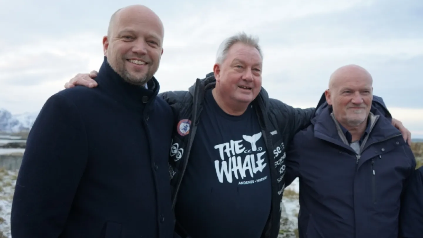 Three adults in dark jackets stand with arms around each other outdoors, with snowy mountains in the distance.