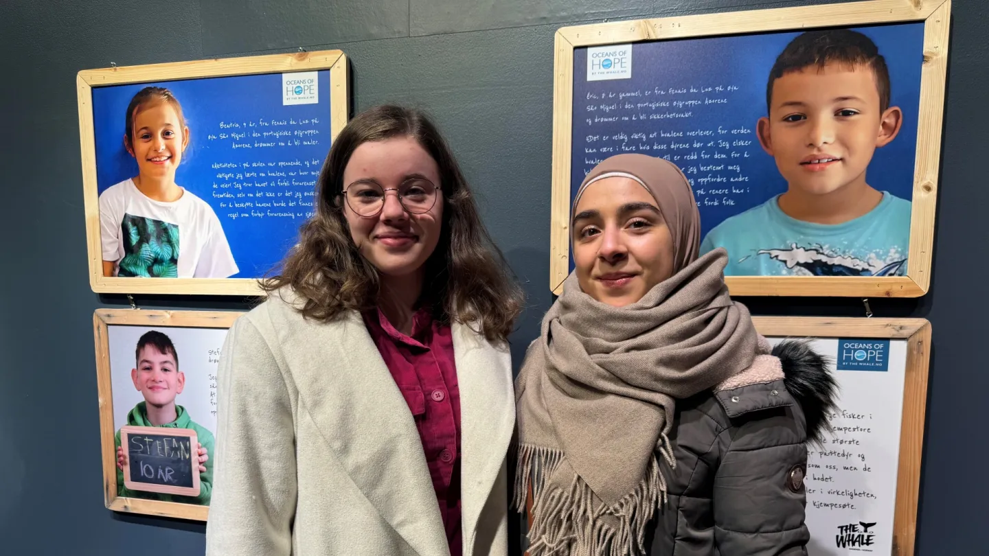 Two people stand indoors in front of framed portrait panels with handwritten text on a dark blue wall.
